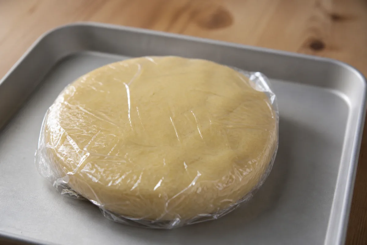 Disque de pâte sablée filmé et aplati sur une plaque, prêt pour le repos au réfrigérateur